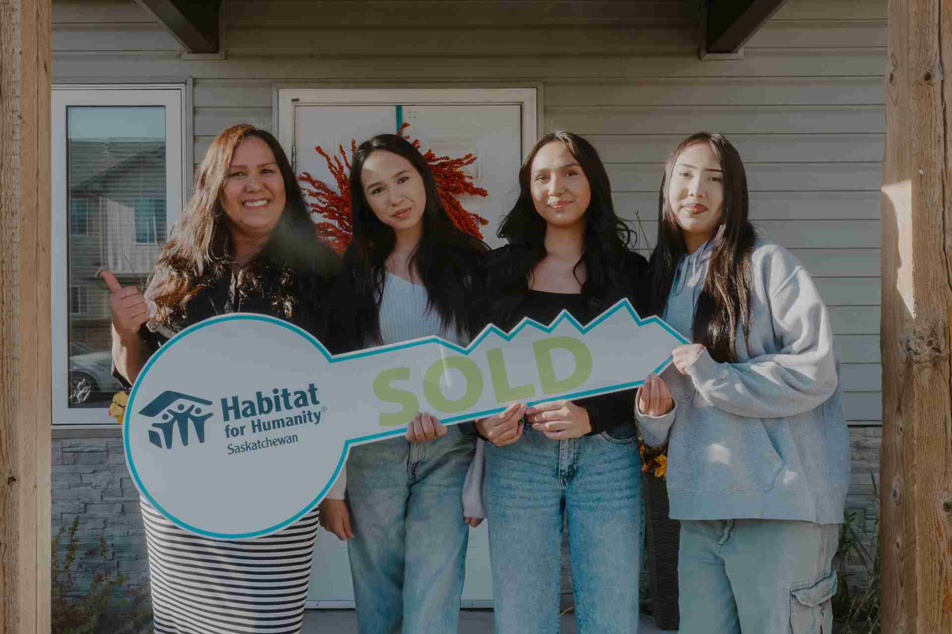 Habitat for Humanity Saskatchewan own a home Yorkton A group of four women, including a Habitat for Humanity representative, are standing in front of a newly purchased home, holding a large "SOLD" key with the Habitat for Humanity Saskatchewan logo. The background features the home’s front door decorated with a red wreath. Everyone is smiling, celebrating the milestone of homeownership.